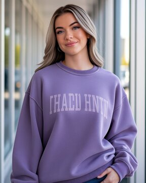 A young woman with blonde hair smiles softly, wearing a purple sweatshirt with white text, standing indoors near large windows with a blurred background.