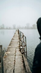 Fototapeta premium Person Standing Near Pier on Calm Lake with Misty Weather and Overcast Sky