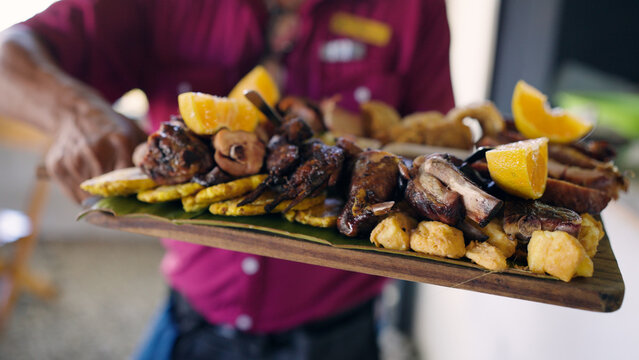 Server holding a large wooden platter with grilled meat, plantains, tostones, and oranges, representing authentic Caribbean cuisine