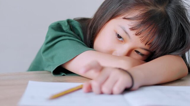 Close up selective focus of demotivated Asian kid girl resting her head on a desk in daylight. She looks overburdened by too much homework, illustrating academic pressure and childhood study burnout.