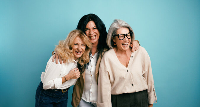 Three diverse mature women friends embracing, smiling, and laughing together with joy and connection on a blue studio background