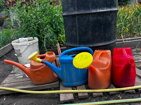 Garden watering cans and plastic containers are arranged near a water barrel. Perfect for horticulture articles, DIY guides, and editorial layouts.