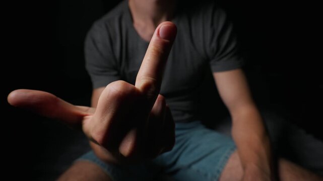 A frustrated young man shows an offensive middle finger gesture directly to the camera in a dimly lit room. Concept of anger, rebellion, aggression, disrespect, and negative attitude.