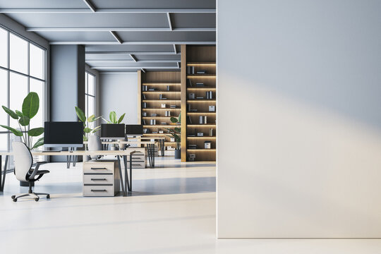 Bright open space office interior with white desks, computer monitors, wooden bookshelves, green plants and empty wall mockup in foreground. 3D Rendering