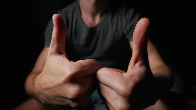 Close-up view shows a man holding two thumbs up directly toward the camera lens in a moody dark setting. Concept of total approval, ultimate success, positive feedback, and complete customer