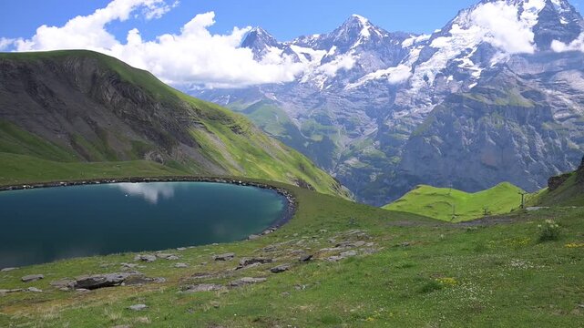 Serene mountain lake and green meadow with dramatic snow-covered peaks and clouds in the Swiss Alps on a clear summer day.