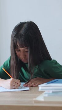 Vertical video showing close up selective focus of a focused asian kid girl writing in a notebook in daylight. Ideal for mobile social media, representing student doing homework and modern education.