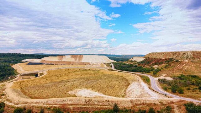 Excavated quarry basin between terraced mineral waste mounds. Winding roads cross a graded depression bordered by pale slopes, grass and a small drainage pond.