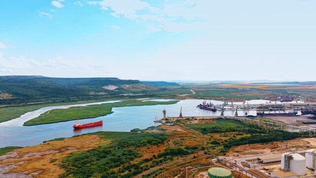 Red tanker sailing past wetlands toward industrial harbor. Elevated view shows a cargo vessel crossing calm water beside marsh islands and dockside cranes.