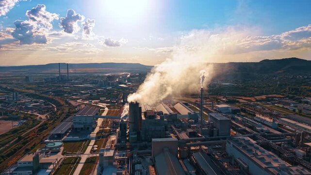 Dense steam drifting above processing factory and pipelines. Backlit emissions rise from multiple stacks over production halls, roads and surrounding hills.