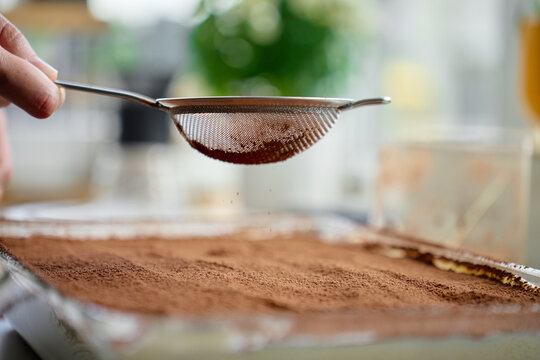 Hand holds sieve with cocoa powder over tiramisu dessert in glass dish