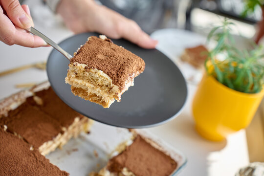 Hand holds tiramisu slice on spatula over black plate, with glass dessert dish in background