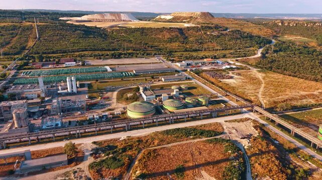 Industrial processing plant with pipelines below terraced quarry. High angle view of green vessels, pipe racks and service roads bordered by wooded hills.
