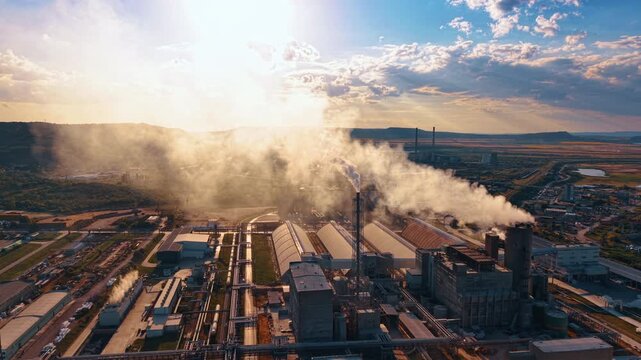 Steam plumes rising above industrial processing buildings at sunset. Backlit vapor crosses factory roofs, vertical stacks and parallel pipes under blue and amber clouds.