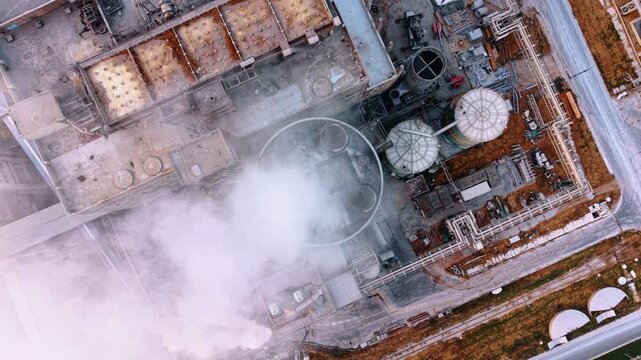 Top view of industrial tower releasing dense steam. Vertical drone perspective shows a circular stack, tanks and pipe grids beneath drifting white vapor.