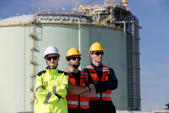 Three engineers in safety gear stand confidently at construction site with industrial storage tanks in background