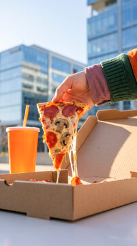 Hand lifting cheesy pepperoni pizza slice from box outdoors in modern city