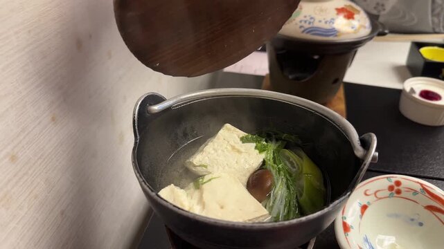 Hand opening a hot nabe pot revealing a steaming traditional Japanese meal with tofu and vegetables