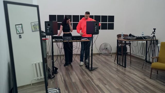 DJ teaches a girl how to mix tracks during his lesson. DJ courses hobby for adults. Music dj producer showing to young woman how to adjust volume on sound mixer in music studio.