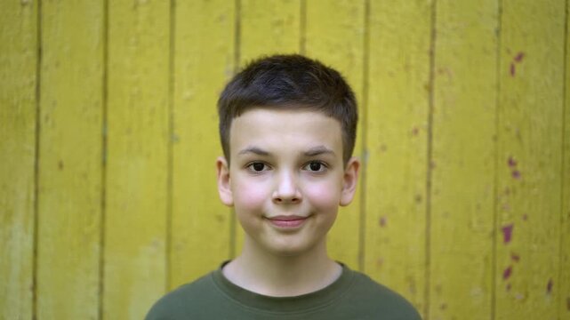 Cute young boy close-up portrait looking at camera with slight smile against yellow wooden wall. Green t-shirt, natural light, innocent expression.