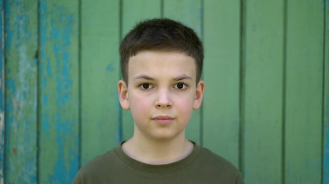 Young boy with short hair in green t-shirt looks seriously at the camera in close-up portrait against green wooden wall with peeling paint.