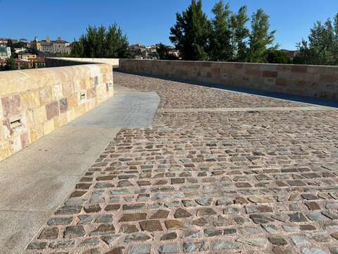 Sunlit stone bridge with a decorative lamp post and cobblestone walkway leading toward an old town skyline beside a calm river. Quiet, historic atmosphere ideal for travel and architecture imagerys