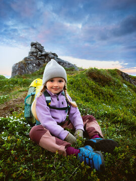 Child girl sitting on grass during hiking trip travel in Norway family vacations outdoor summer tour, 5 years old kid with backpack enjoying wild nature