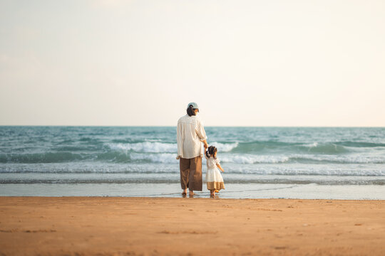Back view of happy Asian mother holding hands with her little daughter while standing on sandy beach, family looking at sea waves during summer vacation for travel tourism, parenting, and childhood