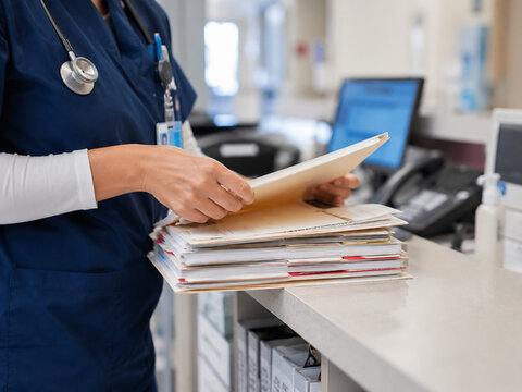 Medical staff member in blue scrubs organizing a stack of patient files and folders at hospital reception desk managing healthcare records and administrative paperwork for clinical database systems.