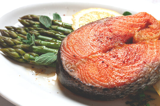 Salmon fillet with asparagus and lemon slices served on a white plate, light marble table, sunlight, home, no people,