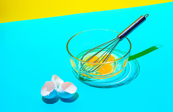 Mixing egg yolks in a glass bowl with a whisk on a bright blue and yellow background in a kitchen setting