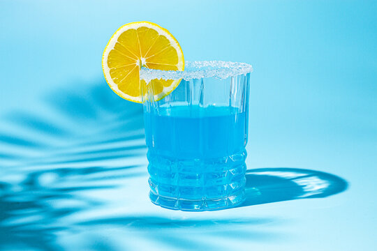 Bright blue drink with lemon slice on a clear glass against a blue background with playful shadows at midday