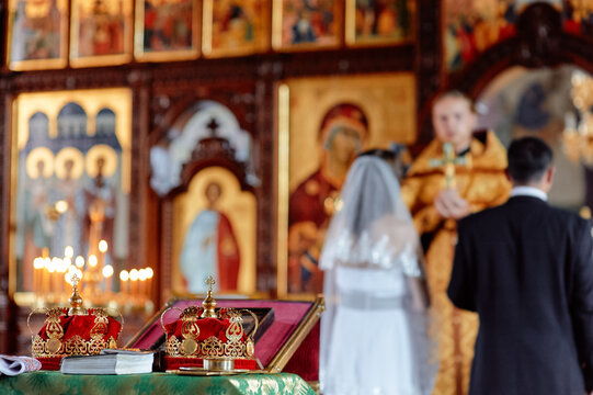 Bride and groom stand before a priest in an ornate Orthodox church as gold wedding crowns rest on the altar; a traditional sacrament filled with faith, ritual, icons and candlelight.