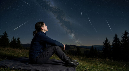 Obraz premium Woman watching the Perseid meteor shower in the night sky. Solitary female sitting on a blanket looking at shooting stars and the Milky Way. Outdoor astronomy and stargazing