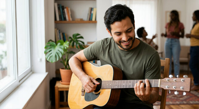 Smiling Hispanic man playing acoustic guitar at home. Happy male musician practicing instrument in living room with friends in background
