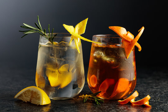A gin and tonic and an Old Fashioned in frosted glasses on a black table.