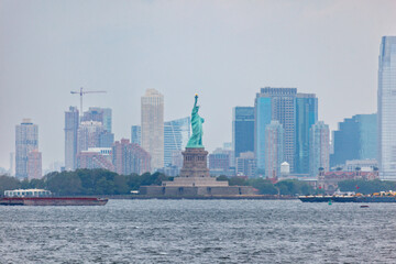 The Statue of Liberty stands tall in New York Harbor, a symbol of freedom and democracy, with the city skyline and passing barges in the foreground. © Pete Niesen Photo