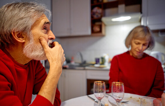 elderly couple sits at kitchen table after dinner, both wearing red sweaters. man looks thoughtful and pensive while the woman appears quiet and reflective, conveying intimacy, fatigue, and distance.