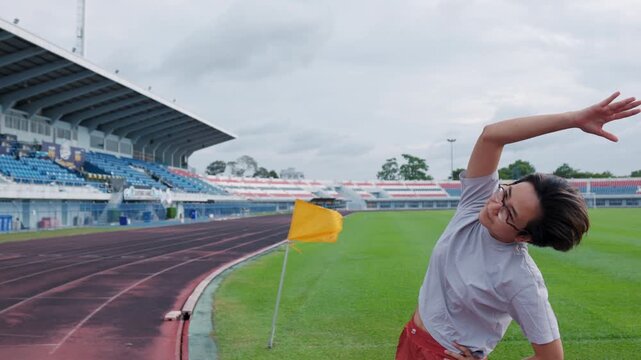 Authentic shot of an active Asian woman in sportswear stretching her arms on a running track. An empty stadium, green soccer field, and waving yellow flag provide excellent copy space.