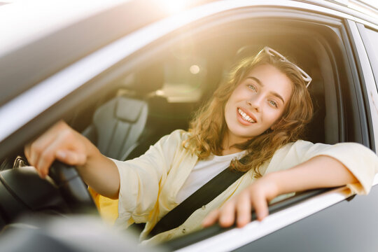 A woman sits behind the wheel of a car, enjoying a sunny day. A smiling female driver sits in the driver's seat and looks out the window. Concepts of transportation, urban mobility, and car rental.