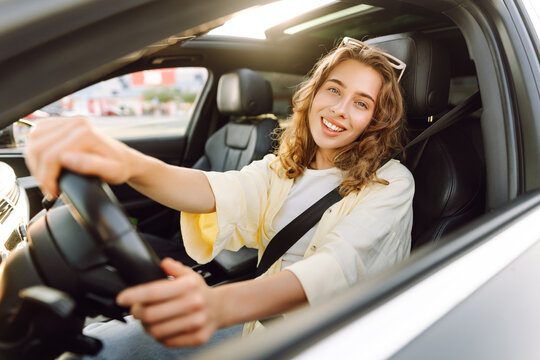 A woman sits behind the wheel of a car, enjoying a sunny day. A smiling female driver sits in the driver's seat and looks out the window. Concepts of transportation, urban mobility, and car rental.