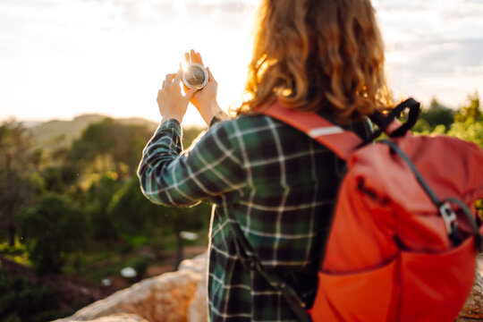 Woman with hiking backpack stands on edge of cliff, holding a compass. View from behind. A young woman hiker is looking for directions while hiking. Concepts: nature, navigation, adventure.