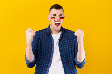 Fototapeta premium Excited Young Caucasian Man With Face Paint Showing Happiness By Raising Fists While Celebrating Sports Event In Bright Yellow Studio Background. Sports Concept.