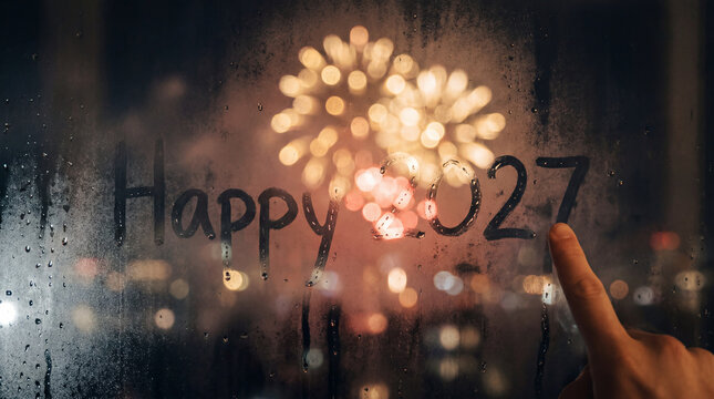 Finger writing Happy 2027 on foggy window with fireworks in background