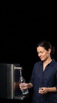 Woman filling reusable water bottle from modern dispenser against black background