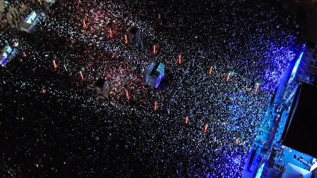 Aerial around view of crowd at night music concert. People gathered at festival outdoor stage with lights and screens, 4k