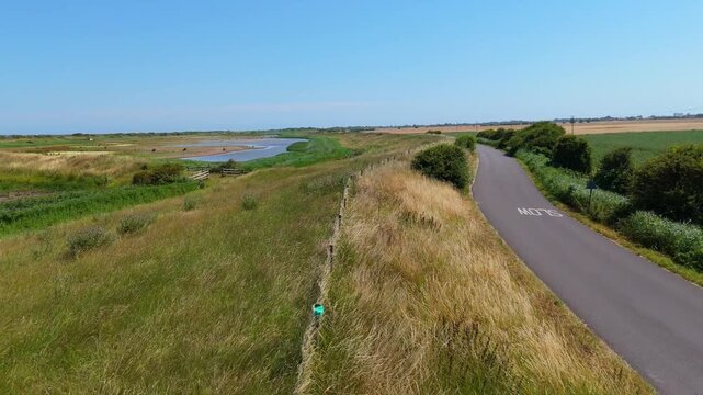 Aerial view shows a quiet countryside path beside a winding river. Grass sways gently in the breeze alongside a paved trail. The sky is clear, and sunlight bathes the natural landscape