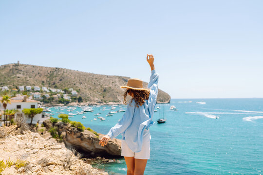 A rear view of a young woman wearing a hat sitting on the edge of a cliff. The woman feels free and enjoys the seascape on a sunny day. Concept of nature, relaxation, and freedom.