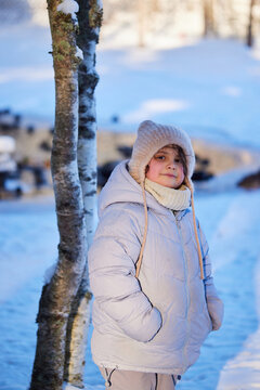 Child stands near tree in snowy field during winter afternoon while wearing warm coat and hat