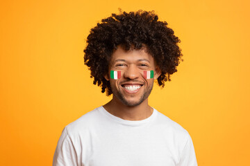 Fototapeta premium Smiling Black Man In His 30s With Italian Flag Face Paint, Enjoying Celebration, Standing Against Bright Yellow Background. Emotion Of Happiness Reflecting Festive Atmosphere, Concept Of Celebration.